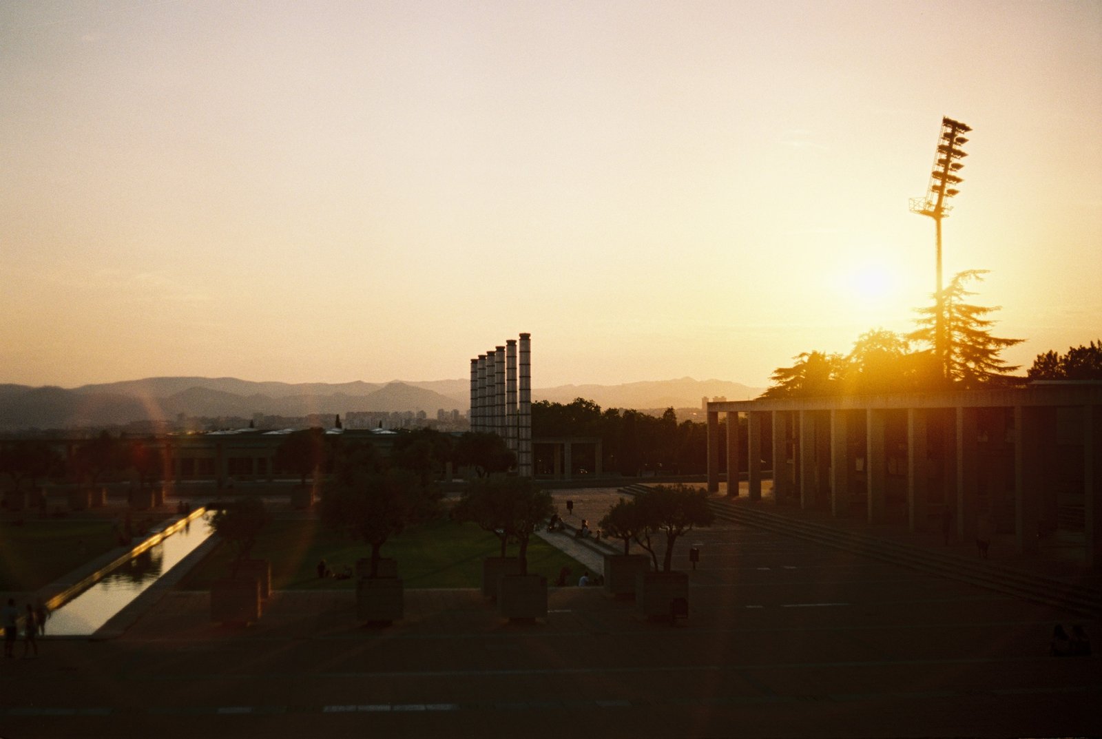 Sunset over a peaceful urban plaza with trees, modern architecture, and mountains in the distance.